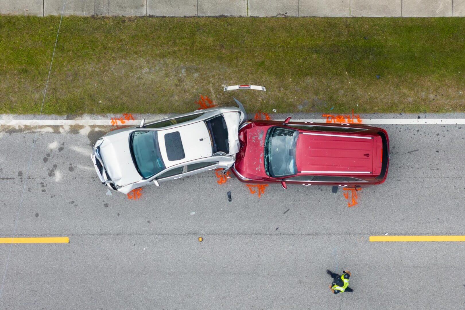 Top view of a car accident involving a white car and a red vehicle on a road. A person in a reflective jacket stands nearby.